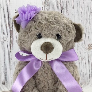 May include: A plush grey teddy bear with a purple flower on its head and a purple ribbon around its neck.