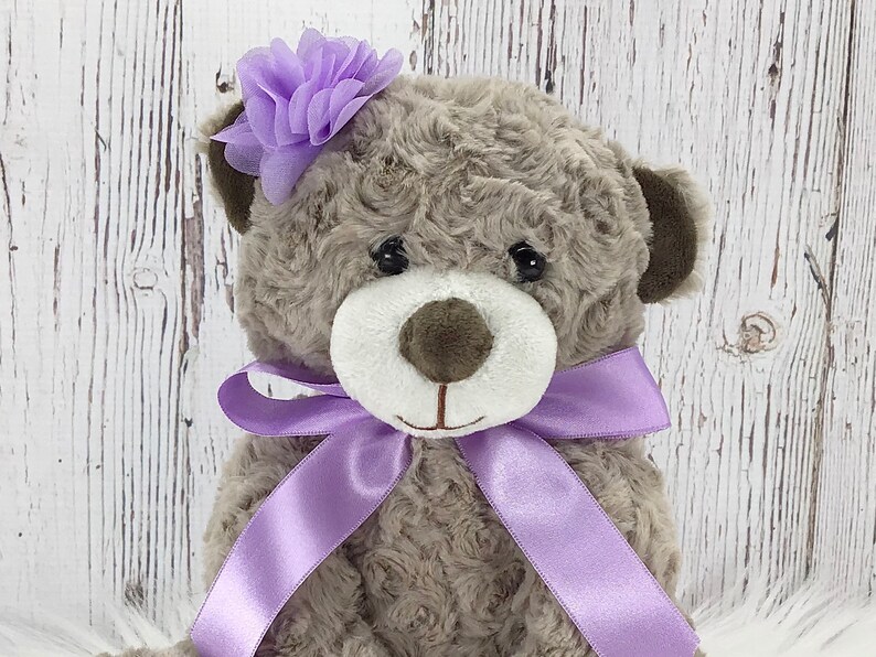 May include: A plush grey teddy bear with a purple flower on its head and a purple ribbon around its neck.