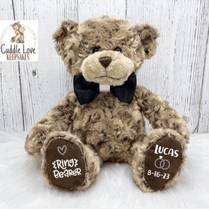 May include: A brown teddy bear wearing a black bow tie. The bear is sitting on a white surface. The bear's paws are embroidered with the text "Ring Bearer" and "Lucas 8-16-23" and a heart and two rings.