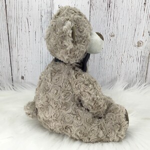May include: A plush grey teddy bear with a black bow tie. The bear has a white face and a fluffy, rosette-like texture.
