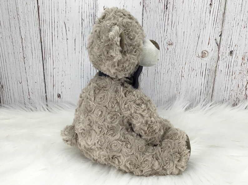 May include: A plush grey teddy bear with a black bow tie. The bear has a white face and a fluffy, rosette-like texture.
