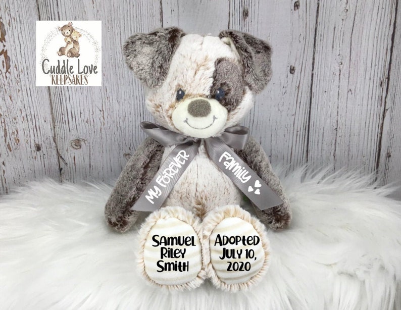 May include: A plush gray and white dog with a gray ribbon that says "My Forever Family". The dog's paws have embroidered text that says "Samuel Riley Smith" and "Adopted July 10, 2020".