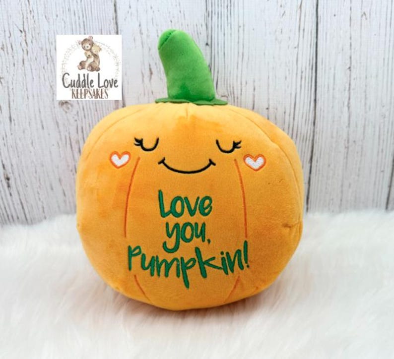 May include: A plush orange pumpkin toy with a green stem, featuring embroidered closed eyes, a smiling mouth, two small white hearts, and the green text "Love you, Pumpkin!" stitched on the front surface of the soft fabric material.