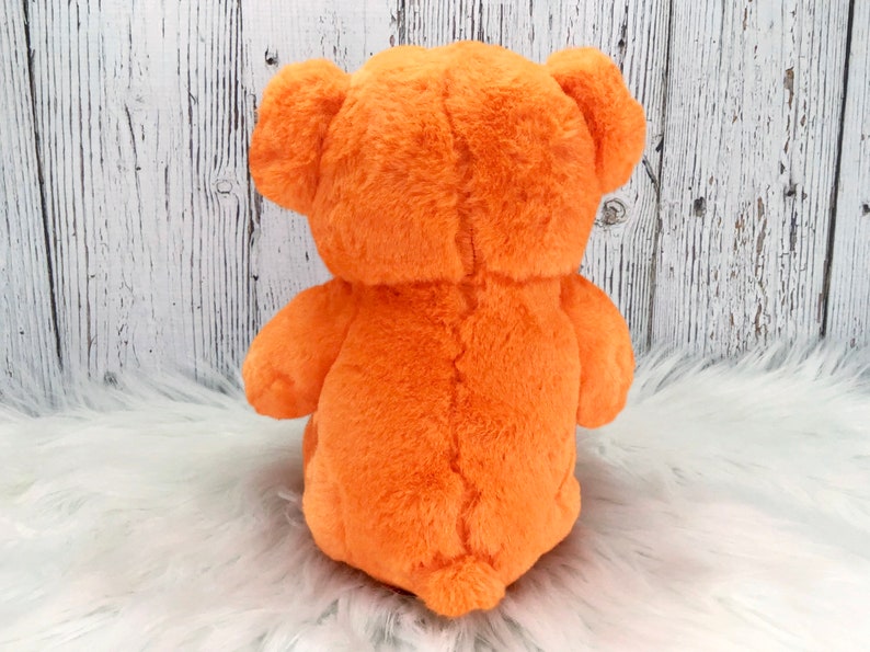 Peut inclure: Vue arri&egrave;re d'un ours en peluche orange. L'ours est assis sur une surface blanche et moelleuse, avec un fond en planches de bois. L'ours a une texture douce et duveteuse.
