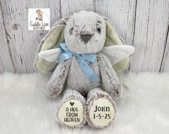 Può includere: Un coniglietto di peluche grigio con ali d'angelo e un fiocco blu. Il coniglietto ha il testo "A HUG FROM HEAVEN" e "John 1-5-25" stampato sui piedi. È appoggiato su una superficie bianca e soffice.