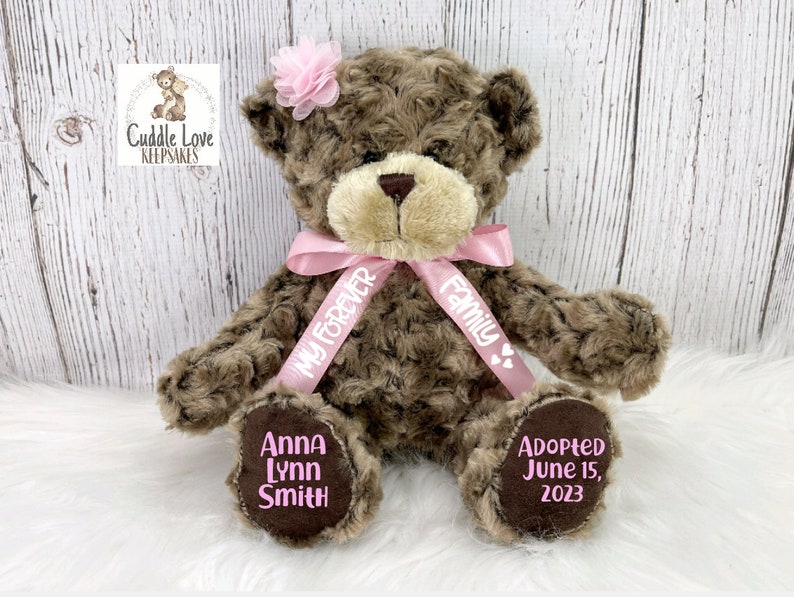 May include: A brown teddy bear with a pink bow and a pink flower on its head. The bear has a pink ribbon around its neck with the text "My Forever Family" on it. The bear's paws have the text "Anna Lynn Smith" and "Adopted June 15, 2023" on them.