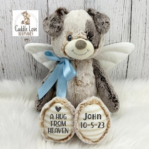 May include: A plush gray and brown dog with white angel wings and a blue ribbon. The dog has a heart on one paw that says "A hug from heaven" and the other paw says "John 10-5-23".