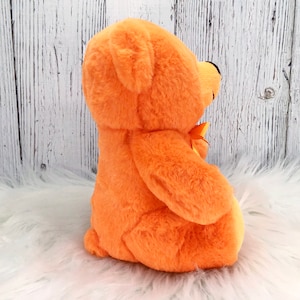 Peut inclure: Un ours en peluche orange avec un petit n&oelig;ud jaune. L'ours a une texture douce et floue et un nez noir. Le fond est une surface blanche textur&eacute;e.