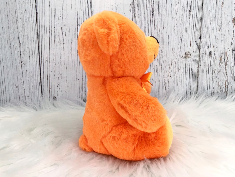Peut inclure: Un ours en peluche orange avec un petit n&oelig;ud jaune. L'ours a une texture douce et floue et un nez noir. Le fond est une surface blanche textur&eacute;e.