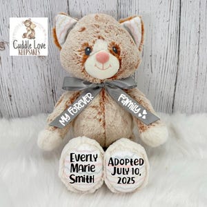 May include: A plush cat-shaped keepsake with brown and white fur, a gray ribbon, and embroidered details. The ribbon reads "My Forever Family." The cat's paws are embroidered with the name "Everly Marie Smith" and the adoption date "July 10, 2025."