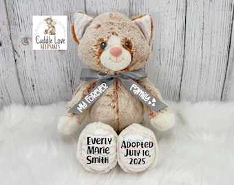 May include: A soft toy cat with brown and white fur, a grey ribbon, and embroidered details. The ribbon reads "My Forever Family." The cat's paws are embroidered with the name "Everly Marie Smith" and the adoption date "July 10, 2025."
