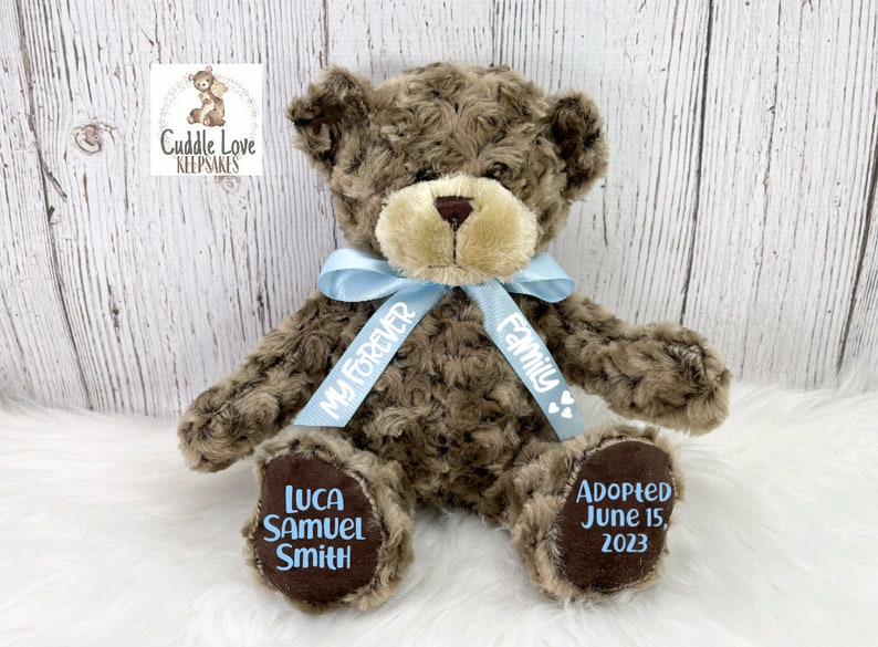 May include: A brown teddy bear with a blue ribbon that says "My Forever Family". The bear's paws are embroidered with the name "Luca Samuel Smith" and the date "Adopted June 15, 2023".