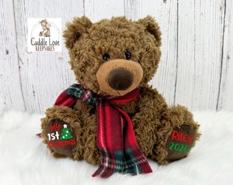 May include: A brown plush teddy bear wearing a red and green plaid scarf. The bear has dark eyes and a brown nose. The left foot pad features the text My 1st Christmas with a tree graphic, and the right foot pad features the name Riley and 2026.