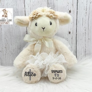 May include: A cream-colored lamb plush toy with a floral crown and a white bow. The lamb has the name "Alice" and a cross printed on one foot, and "Baptized 9-8-26" with footprints on the other. The lamb is wearing a white tulle skirt.