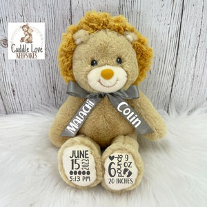 May include: A plush lion teddy bear with a gray ribbon tied around its neck. The ribbon has the names "Malachi" and "Colin" printed on it. The bear's paws have details printed on them, including the date "June 15, 2023", the time "5:13 PM", the weight "6 pounds 9 oz", and the length "20 inches".