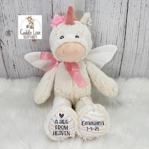 Pode incluir: Um brinquedo de pel&uacute;cia de unic&oacute;rnio creme com uma fita, chifre e flor rosa. O unic&oacute;rnio tem asas brancas e o texto "A HUG FROM HEAVEN" e "GRANDMA 1-5-25" nos p&eacute;s. O logotipo "Cuddle Love Keepsakes" est&aacute; no canto superior esquerdo.