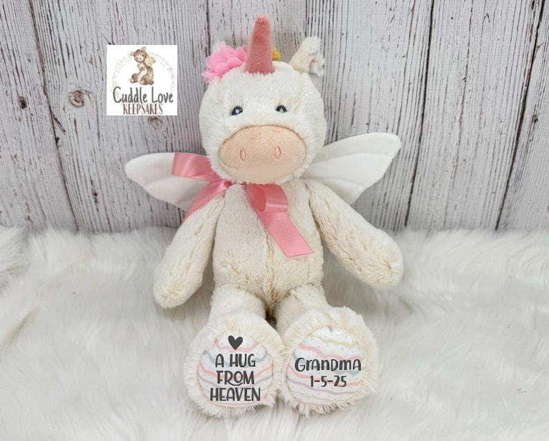 Pode incluir: Um brinquedo de pel&uacute;cia de unic&oacute;rnio creme com uma fita, chifre e flor rosa. O unic&oacute;rnio tem asas brancas e o texto "A HUG FROM HEAVEN" e "GRANDMA 1-5-25" nos p&eacute;s. O logotipo "Cuddle Love Keepsakes" est&aacute; no canto superior esquerdo.