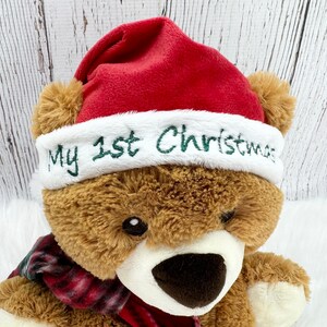 May include: A brown plush teddy bear wearing a red Santa hat with a white plush brim. The white brim features the embroidered green text My 1st Christmas. The bear also wears a red and black plaid scarf around its neck, with a white muzzle.