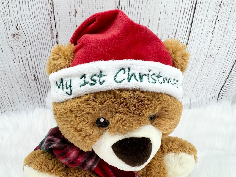 May include: A brown plush teddy bear wearing a red Santa hat with a white plush brim. The white brim features the embroidered green text My 1st Christmas. The bear also wears a red and black plaid scarf around its neck, with a white muzzle.