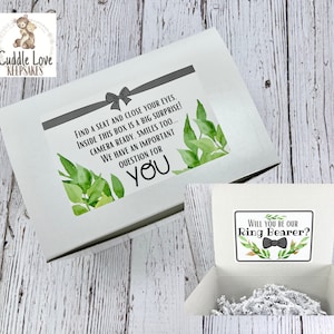 May include: A white box with a green leafy design and the text "Find a seat and close your eyes. Inside this box is a big surprise! Camera ready. Smiles too... We have an important question for you." The box is open and inside is a white card with the text "Will you be our Ring Bearer?" with a black bow tie and green leafy design.