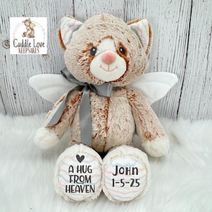 May include: A plush cat with angel wings, a gray ribbon, and the text "A HUG FROM HEAVEN" and "John 1-5-25" on its paws. The cat is primarily brown and white, with a pink nose and a smiling face. The logo "Cuddle Love Keepsakes" is in the background.
