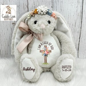 May include: A plush gray bunny with long floppy ears, wearing a floral headband. The bunny has a cross with flowers on its chest that says "I am a child of God". The bunny's paws are embroidered with the names "Ashley" and "Baptized 5-25-24".