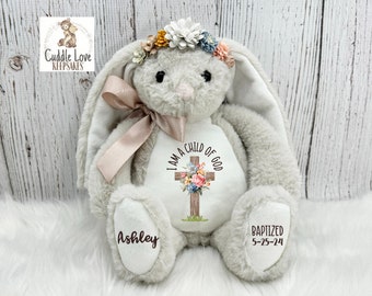 Puede incluir: Un conejito de peluche gris con orejas largas y caídas, que lleva una diadema floral. El conejito tiene una cruz con flores en el pecho que dice "Soy un hijo de Dios". Las patas del conejito están bordadas con los nombres "Ashley" y "Bautizado 5-25-24".