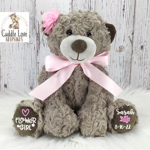 May include: A brown teddy bear with a pink bow and a pink flower on its head. The bear has a pink heart and the words "Flower Girl" embroidered on one paw and the name "Sarah" and the date "8-16-22" embroidered on the other paw.