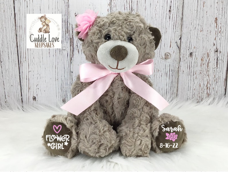 May include: A brown teddy bear with a pink bow and a pink flower on its head. The bear has a pink heart and the words "Flower Girl" embroidered on one paw and the name "Sarah" and the date "8-16-22" embroidered on the other paw.