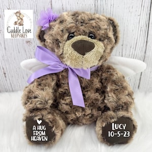 May include: A brown teddy bear with a light brown muzzle, wearing a purple bow and a matching flower. The bear has white angel wings and the text "A HUG FROM HEAVEN" and "LUCY 10-5-23" on its paws. The bear is a keepsake.