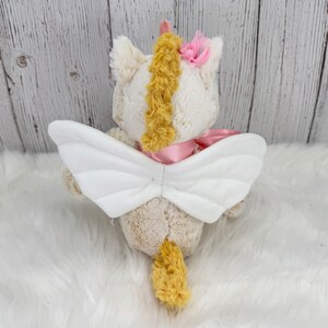 Pode incluir: Um brinquedo de unic&oacute;rnio de pel&uacute;cia com asas brancas, uma fita rosa e uma crina e cauda amarelas. O unic&oacute;rnio tem uma flor rosa na cabe&ccedil;a e um chifre amarelo. O brinquedo est&aacute; em uma superf&iacute;cie branca e fofa.