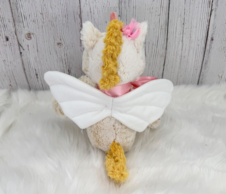 Pode incluir: Um brinquedo de unic&oacute;rnio de pel&uacute;cia com asas brancas, uma fita rosa e uma crina e cauda amarelas. O unic&oacute;rnio tem uma flor rosa na cabe&ccedil;a e um chifre amarelo. O brinquedo est&aacute; em uma superf&iacute;cie branca e fofa.