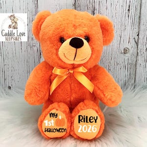 Peut inclure: Un ours en peluche orange avec un n&oelig;ud jaune, des yeux noirs et un visage souriant. Les pieds de l'ours portent l'inscription "my 1st Halloween" et "Riley 2026". L'ours est assis sur une surface blanche et duveteuse.