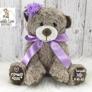 May include: A brown teddy bear with a purple bow and flower headband. The bear has a heart with the words "Flower Girl" on one paw and the name "Sarah" and the date "8-16-22" on the other paw.