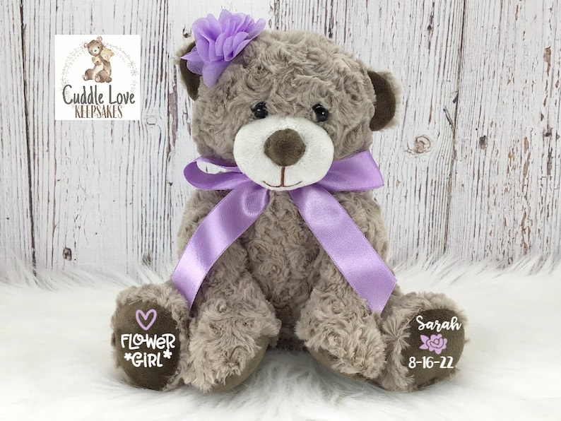 May include: A brown teddy bear with a purple bow and flower headband. The bear has a heart with the words "Flower Girl" on one paw and the name "Sarah" and the date "8-16-22" on the other paw.