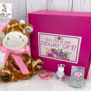 May include: Pink gift box with a giraffe plush toy, a lollipop, a bottle of bubbles, and a necklace with a flower girl charm. The box has a label that says "Will you be our Flower Girl?"