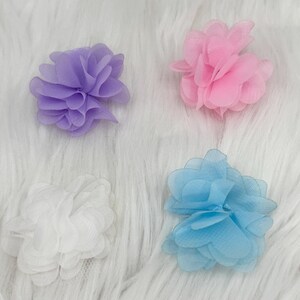 Pode incluir: Quatro enfeites de flores de tecido em cores pastel: lavanda, rosa, branco e azul claro. Cada flor tem m&uacute;ltiplas camadas de p&eacute;talas, criando um efeito texturizado. As flores est&atilde;o dispostas sobre uma superf&iacute;cie branca e fofa.