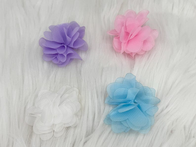 Pode incluir: Quatro enfeites de flores de tecido em cores pastel: lavanda, rosa, branco e azul claro. Cada flor tem m&uacute;ltiplas camadas de p&eacute;talas, criando um efeito texturizado. As flores est&atilde;o dispostas sobre uma superf&iacute;cie branca e fofa.