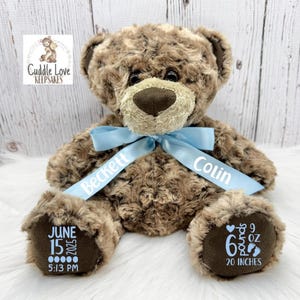 May include: A brown teddy bear with a light blue ribbon. The ribbon is printed with the names "Beckett" and "Colin". The bear's paws are printed with the birth date, time, weight (9 oz), and length (20 inches). The bear has a brown nose and a logo that says "Cuddle Love Keepsakes".