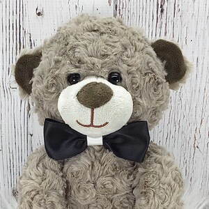 May include: A grey teddy bear wearing a black bow tie. The bear has brown ears and black eyes. The bear is sitting on a white surface.