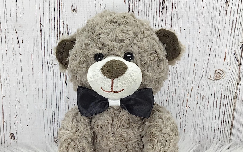 May include: A grey teddy bear wearing a black bow tie. The bear has brown ears and black eyes. The bear is sitting on a white surface.