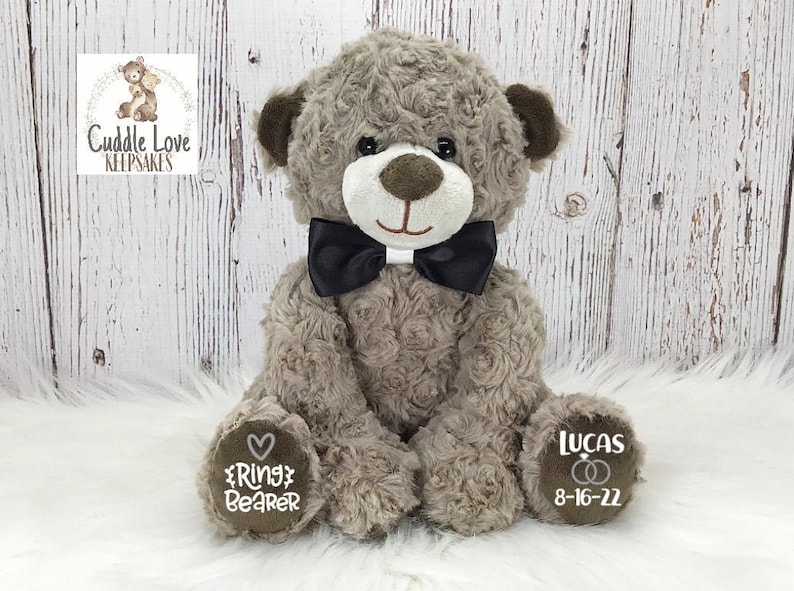May include: A grey teddy bear wearing a black bow tie and holding a sign that says "Ring Bearer". The bear's other paw has a sign that says "Lucas 8-16-22".