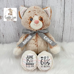 Peut inclure: Une peluche en forme de chat de couleur beige et blanc avec un ruban gris. Le ruban porte les noms "Abigail" et "Grace". Les pattes du chat affichent les détails de la naissance : 15 juin 2025, 17h13, 280 g, 2,7 kg et 51 cm.