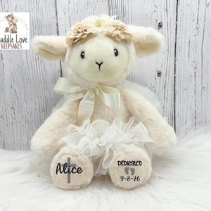 May include: A plush cream-colored lamb stuffed animal wearing a white tulle skirt and a matching ribbon around its neck. The lamb has a floral headband and personalized text on its feet reading Alice with a cross and Dedicated 9-8-26.