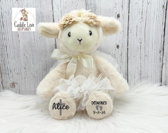 May include: A plush cream-colored lamb stuffed animal wearing a white tulle skirt and a matching ribbon around its neck. The lamb has a floral headband and personalized text on its feet reading Alice with a cross and Dedicated 9-8-26.