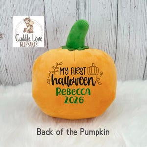 May include: A plush orange pumpkin toy with a green stem. The front features black embroidered text reading My First Halloween with a small pumpkin icon, and the name Rebecca in green text above the year 2026 in green text on the pumpkin surface.
