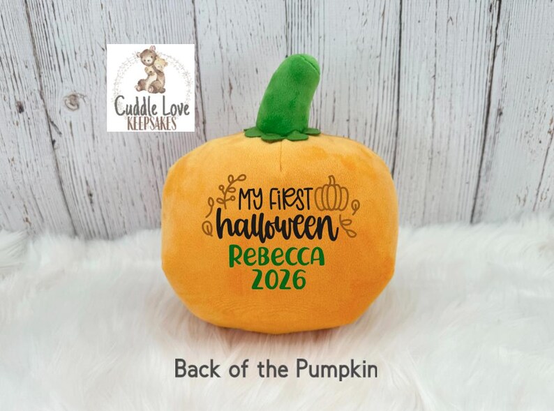 May include: A plush orange pumpkin toy with a green stem. The front features black embroidered text reading My First Halloween with a small pumpkin icon, and the name Rebecca in green text above the year 2026 in green text on the pumpkin surface.