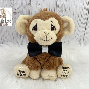 May include: A brown plush monkey wearing a black bow tie with a white stripe. The monkey is sitting on a white fluffy surface. The monkey's left foot has the text "Ring Bearer" and the right foot has the text "Lucas 8-16-24".