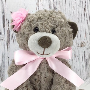 May include: A brown teddy bear with a pink bow and a pink flower on its head. The bear is sitting on a white wooden background.