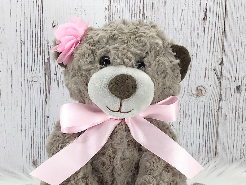 May include: A brown teddy bear with a pink bow and a pink flower on its head. The bear is sitting on a white wooden background.